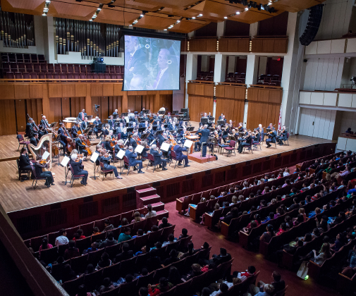 The National Symphony Orchestra performing on the Kennedy Center Concert Hall stage with a small portion of the audience shown.