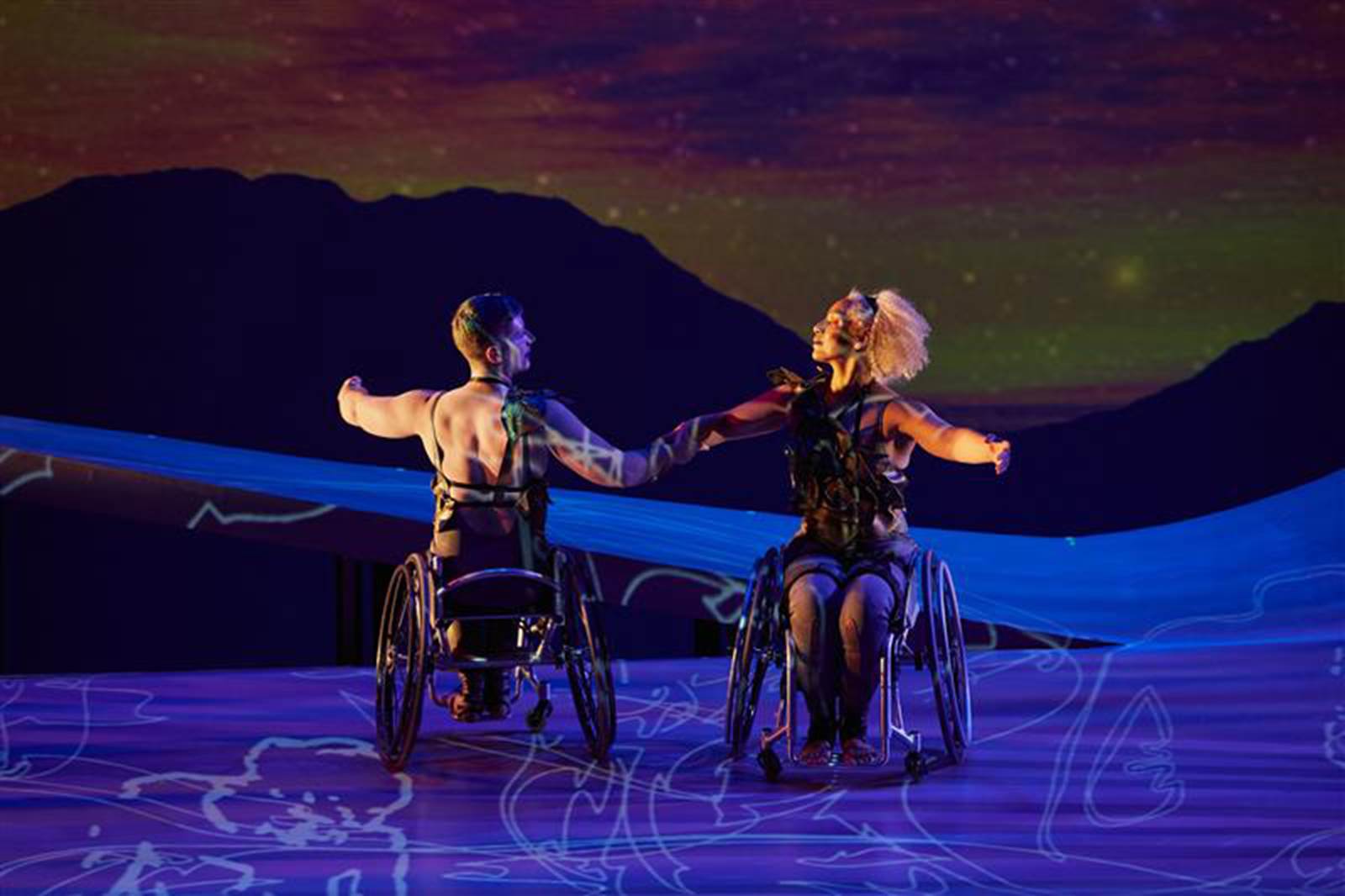 Laurel Lawson and Alice Sheppard hold each others’ forearms, chins lifted as they extend the other curved arm to the side, in a counterbalance turn. Alice is a Black multiracial woman with blonde curly hair and coffee colored skin; Laurel is a white person with cropped teal hair. They both wear gray leggings and leatherlike sleeveless tops that resemble armor or petals. Projected illustrated figures dance across the purpleish stage as a mountain range appears, silhouetted in the sunset, in the background. Photo Jaqlin Medlock / Rutgers University.