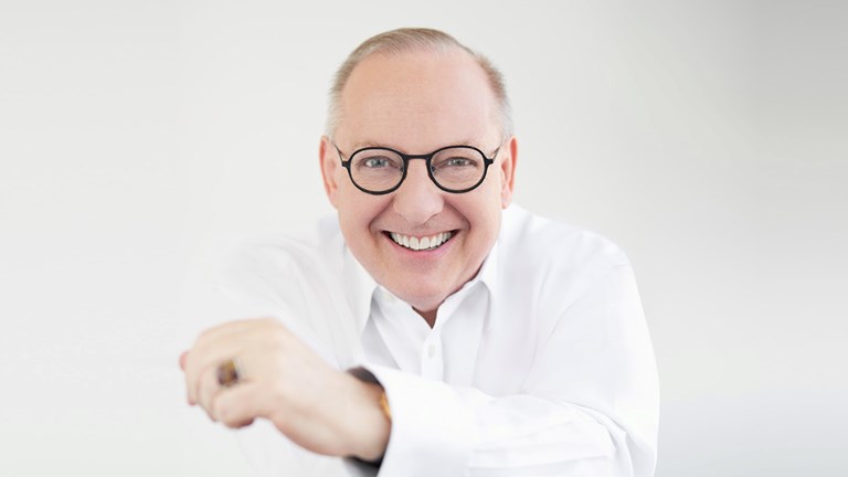 Nicholas McGegan smiling with dark rimmed glasses and semi-crossed arms with his left arm in the forefront