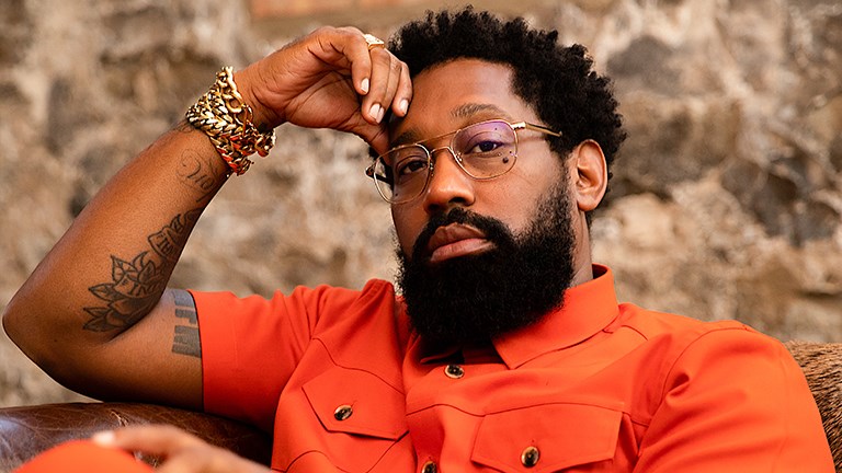 PJ Morton sitting on a leather cofa waering an orange shirt.