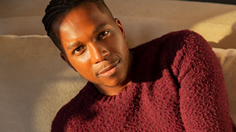 Leslie Odom, Jr. in a textured maroon sweater resting on a kaki couch