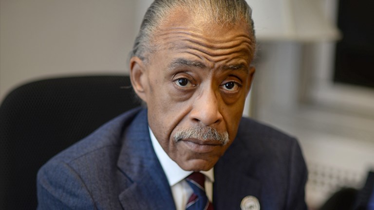 Rev. Dr. Al Shaprton sitting at a desk wearing a navy pinstriped suit
