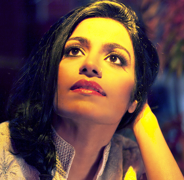 A woman with long black hair and wearing a white blouse gazes into a light above and to the left of the camera. She is leaning the back of her head on her left hand. Her elbow is resting on a table.
