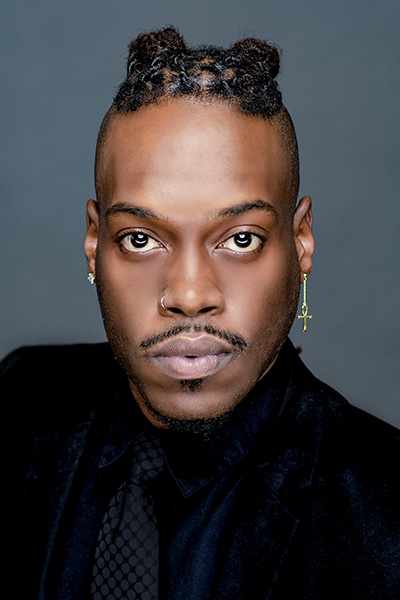 A headshot of singer Daniel Smith who wears a dark shirt and tie in front of a blueish-gray background.