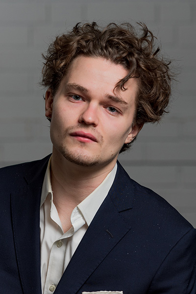 A headshot of Dennis Chemelensky in which he wears a dark jacket with a white dress shirt and is front of a light-colored brick background.