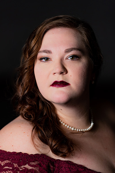 A headshot of singer Suzannah Waddington in which she wears a dark red lacey top with matching lipstick and a white pearl necklace.