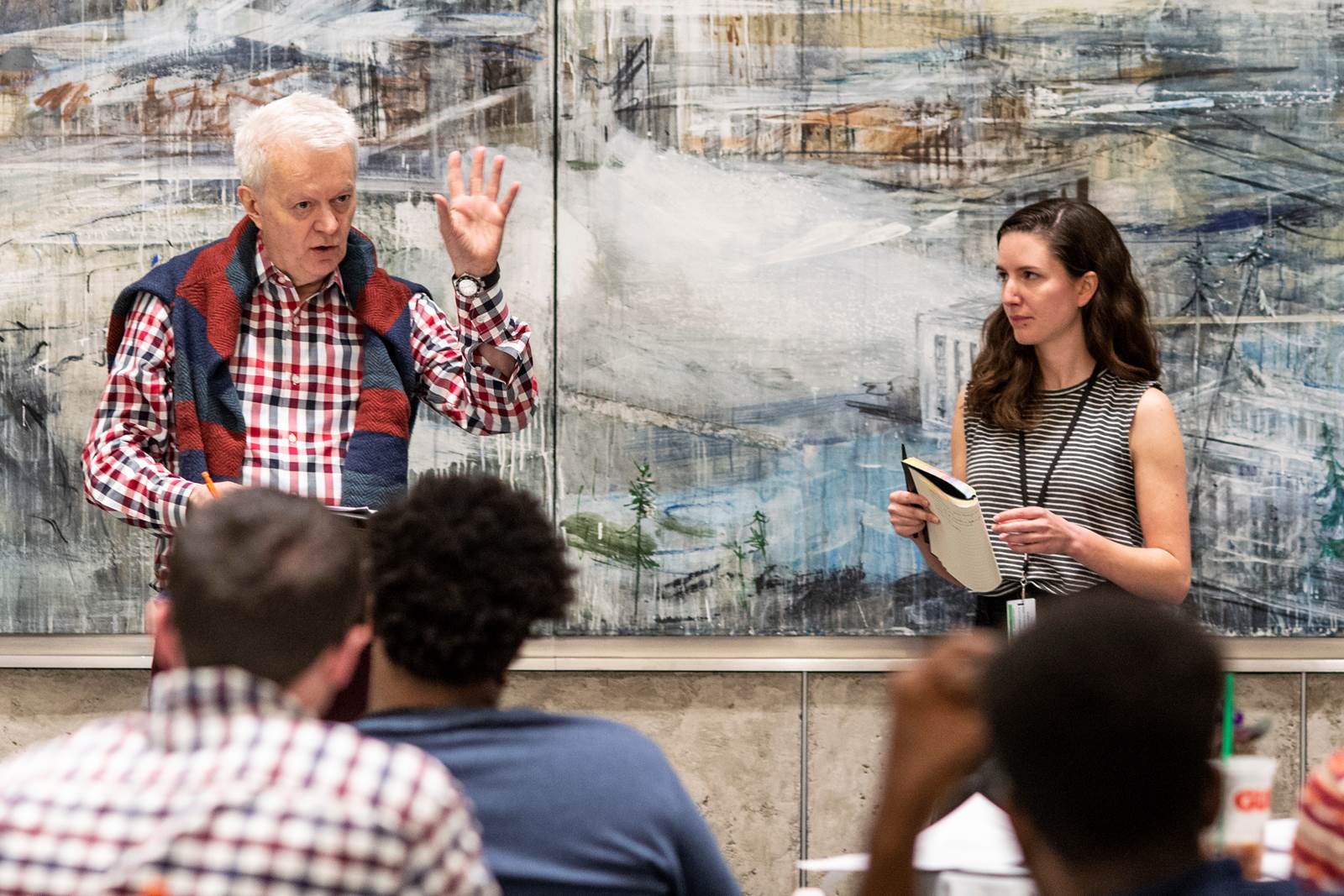 A female student stands beside an instructor as he speaks to the class. There is a large oil painting behind them. 