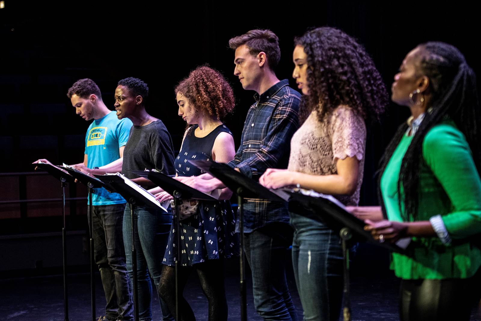 Six actors stand in a row, at music stands.