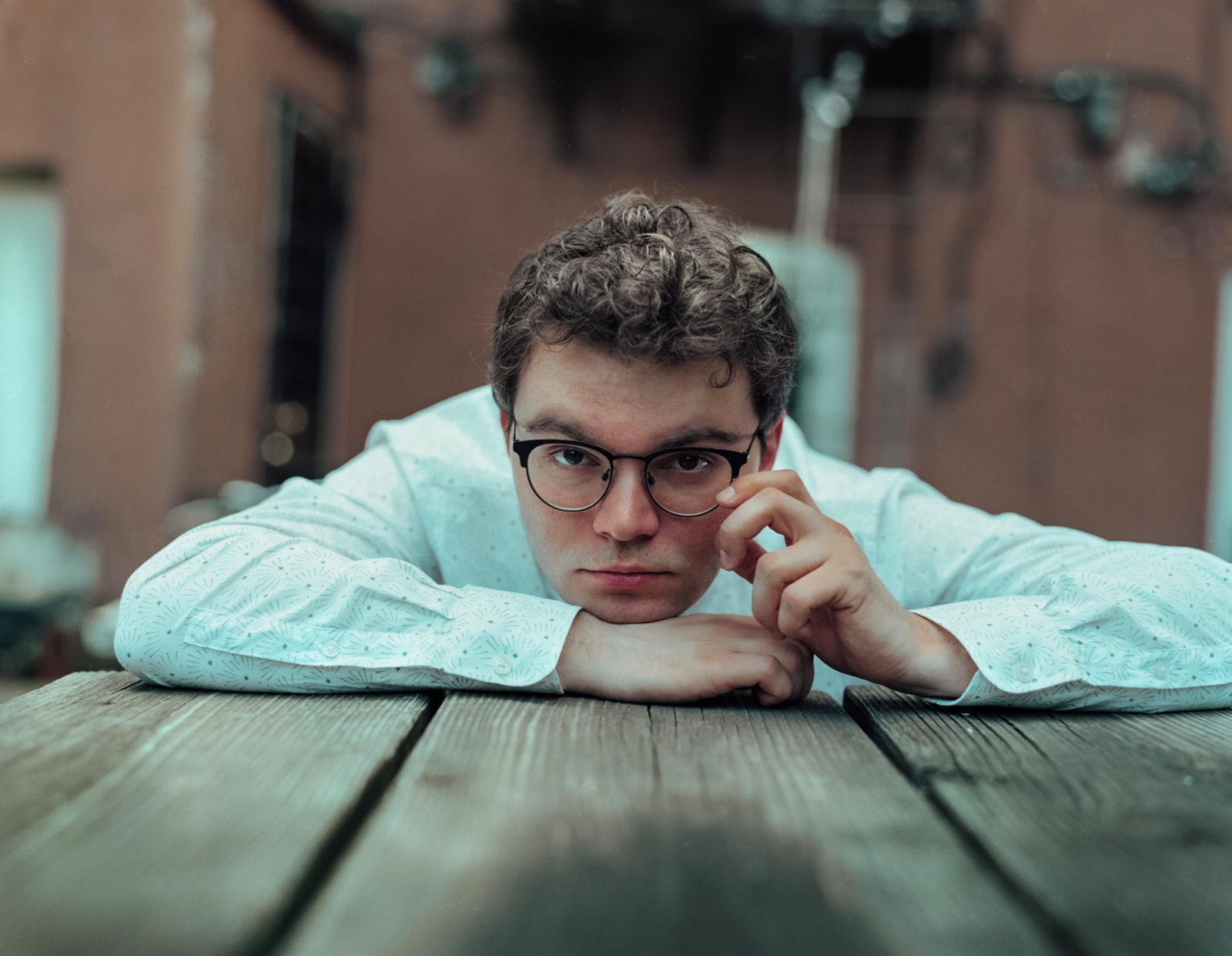 A white person with short curly hair and black-framed glasses wearing a long-sleeve white button-down shirt is leaning on a picnic table looking at the camera with a serious expression.