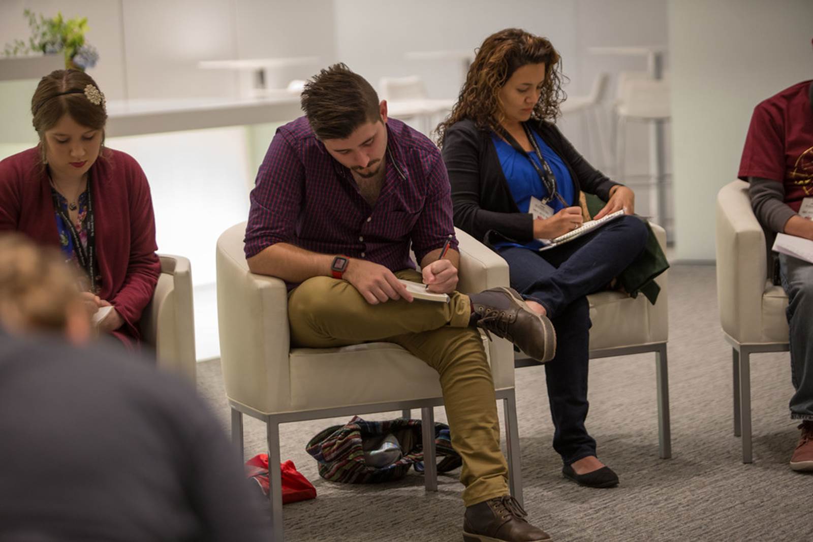 Three students in white armchairs write on notebooks, which they hold in their laps.