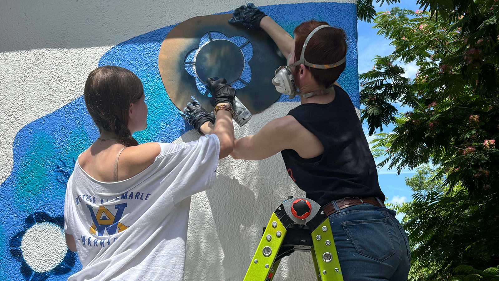 A young artist assists Laura Lee up on a ladder spray painting a stenciled flower design.