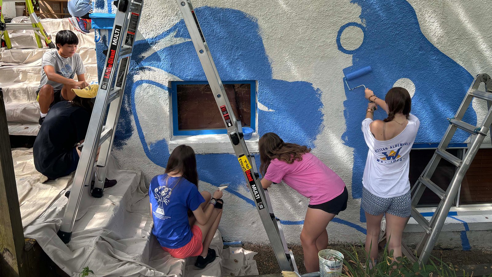 Six young artists are working on the beginning of a mural outdoors on the wall of a building, painting among dropcloths and ladders with rollers and brushes.