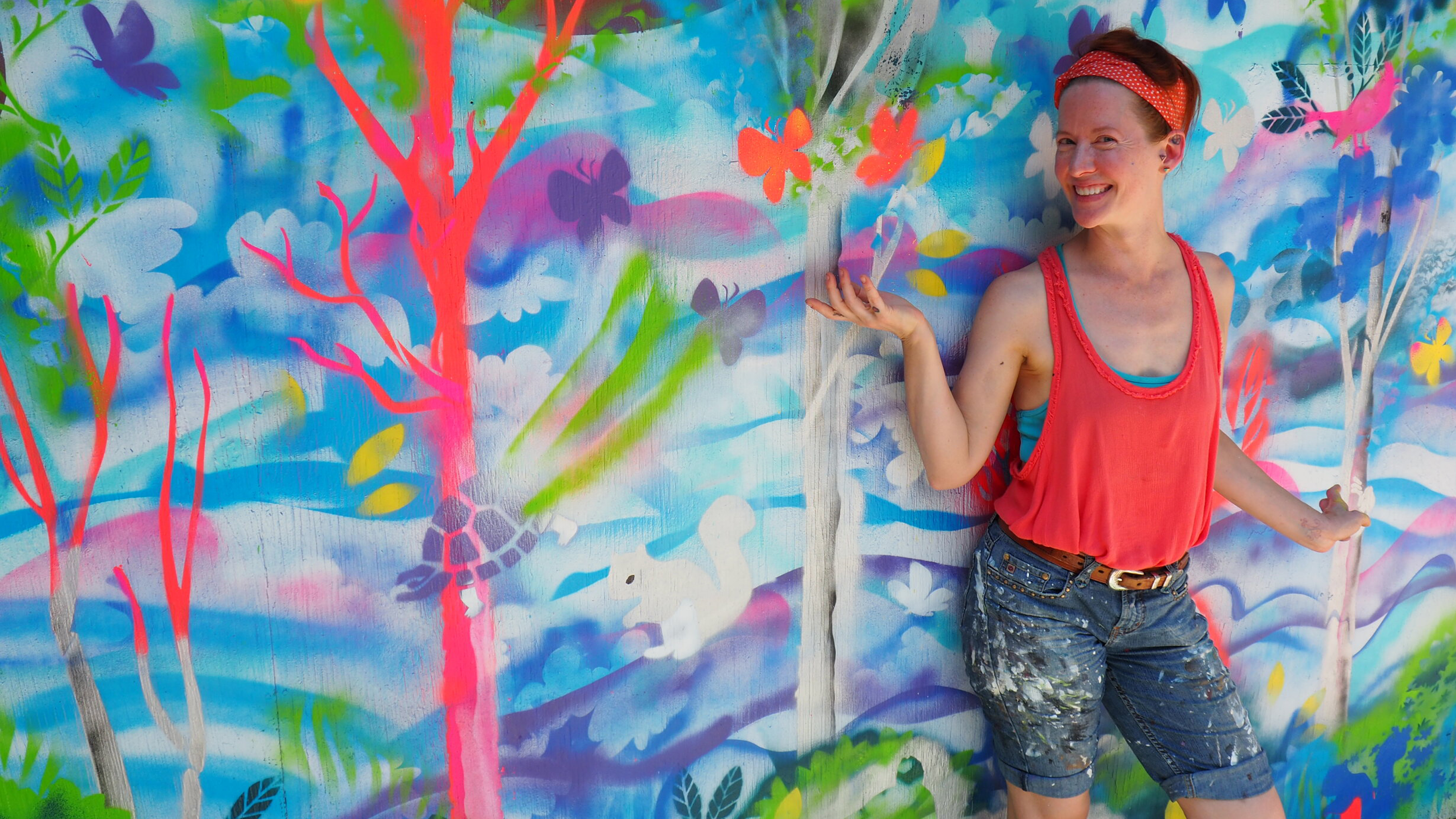 Laura Lee Gulledge, a white woman with short red hair wearing an organge headband, tanktop, and paint-spattered denim shorts is standing in front of a colorful mural of animals and trees.