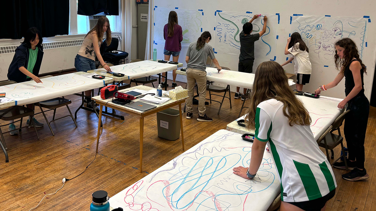 Eight young artists in a classroom drawing on large sheets of paper and hanging them on the walls.