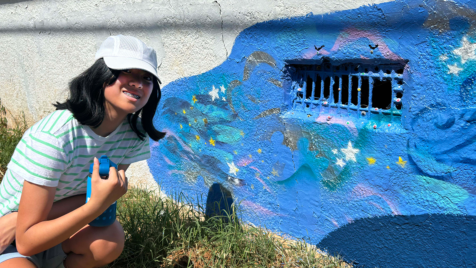 A young artist with long dark hair, a white baseball cap and braces is crouched next to a section of a mural that has incorporated a metal grate in the wall as a fun monster mouth.