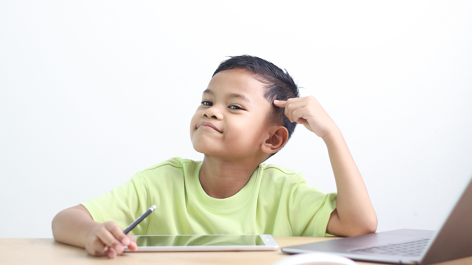 Photo of a young boy with short brown hair wearing a light green t-shirt thinking while holding a stylus, sitting at a desk with a tablet and laptop.