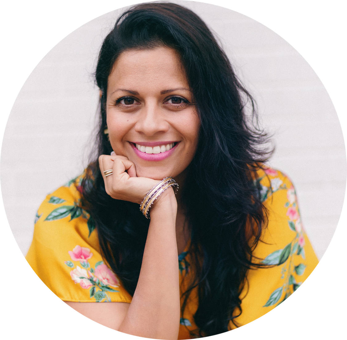 A headshot of Rupa Mehta. She is wearing a yellow dress with a floral design and her black hair is long with waves. Her fist is resting on her chin as she smiles at the camera.