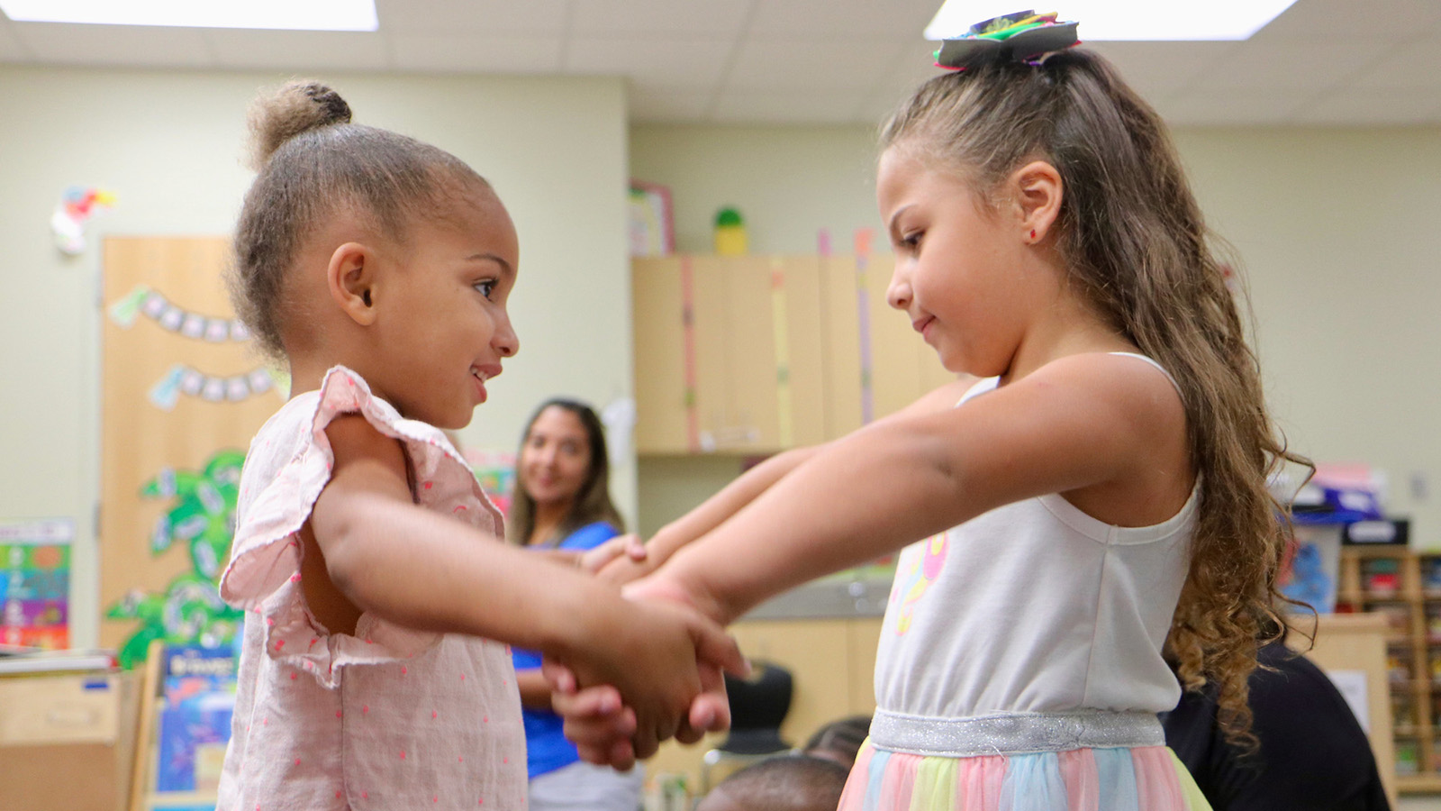 Two young girls in a classroom. They are facing each other and holding hands. 