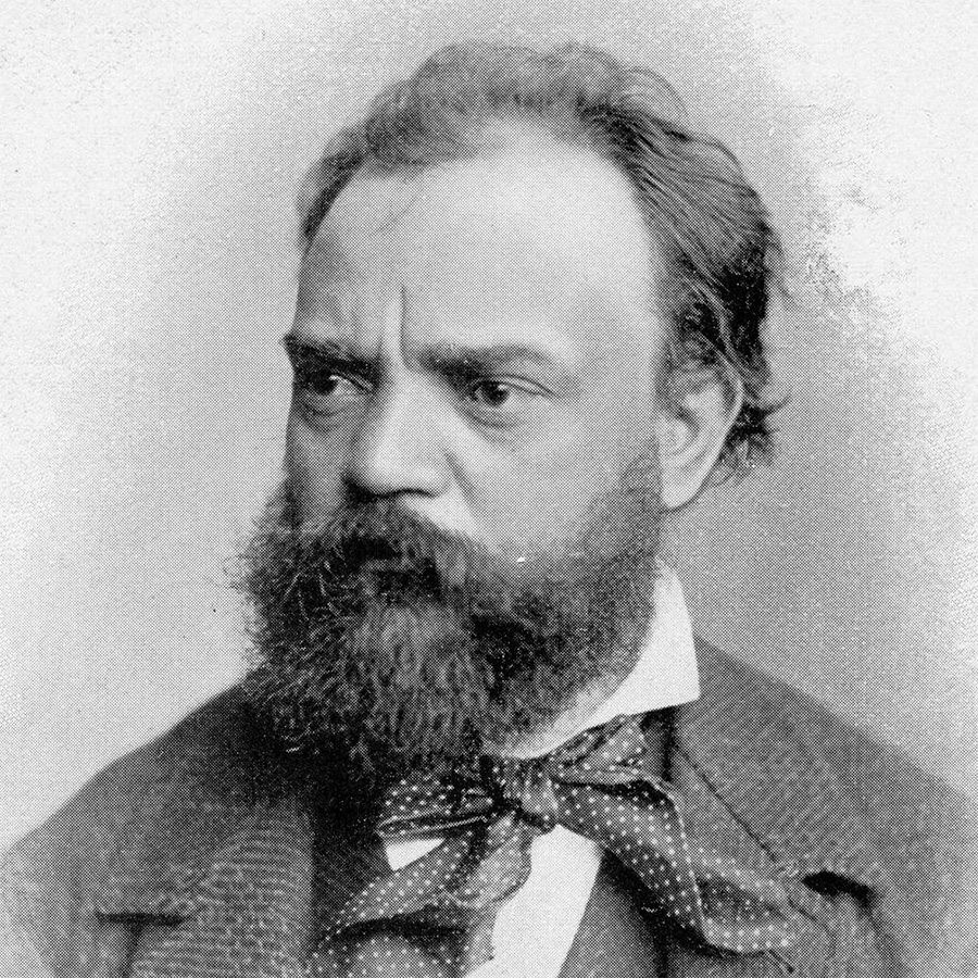 A black-and-white vintage image of Czech composer Antonin Dvorak. He is a white man with a thick beard and dark hair. He wears a dark jacket with a light stripe pattern, a matching vest, and a white dress shirt. He also wears a polka-dotted cravat or scarf tied into a bow underneath his dress shirt&rsquo;s collar.