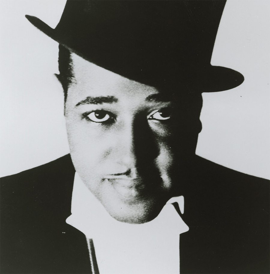 A black-and-white photo of composer, jazz musician, and bandleader Duke Ellington. He wears a slightly tilted black top hat, black jacket, and white dress shirt.  