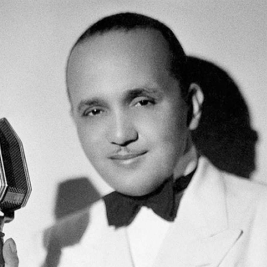 A black-and-white photo of jazz bandleader, arranger, and pianist Fletcher Henderson. He wears a white suit with a black bowtie and holds the stand to an old school microphone.
