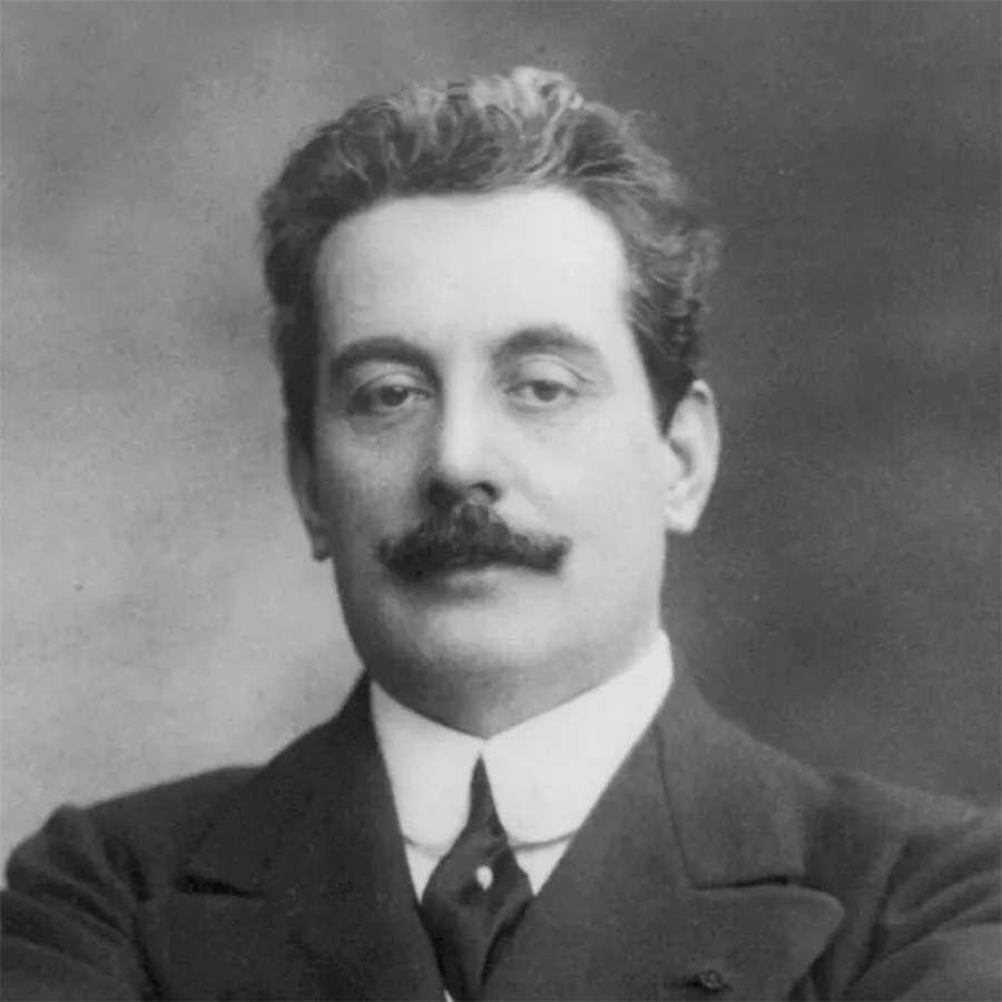 A black-and-white photo of opera composer Giacomo Puccini. He has short dark hair with a mustache. He wears a dark jacket with a white dress shirt and dark tie.
