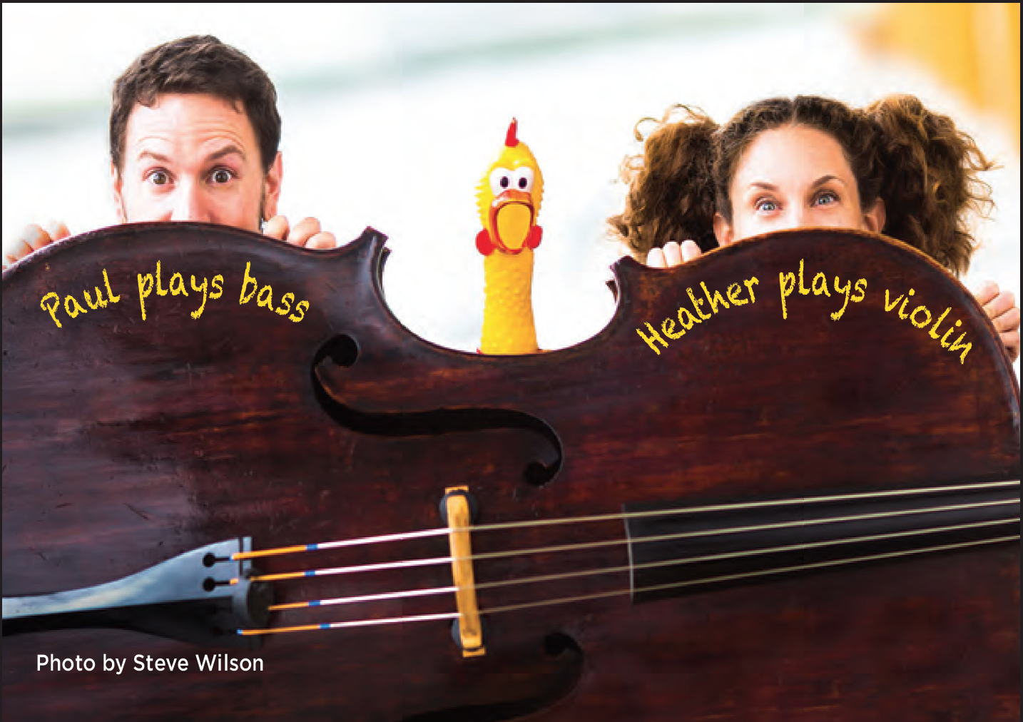 The tops of two people's heads, one with short brown hair, the other with long brown hair in pigtails peeking up from behind a double bass with the head of a rubber chicken peeking out between them.