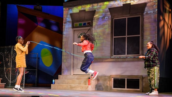 Three girls on stage in front of the She A Gem set playing a Double-Dutch jumprope game. Two of the girls are holding the ends of the rope and the third actor is jumping in the middle.