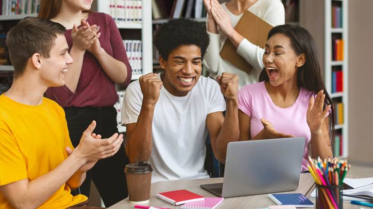 A student looking at a computer with an excited expression. The student is surrounded by four classmates who are cheering and clapping. 