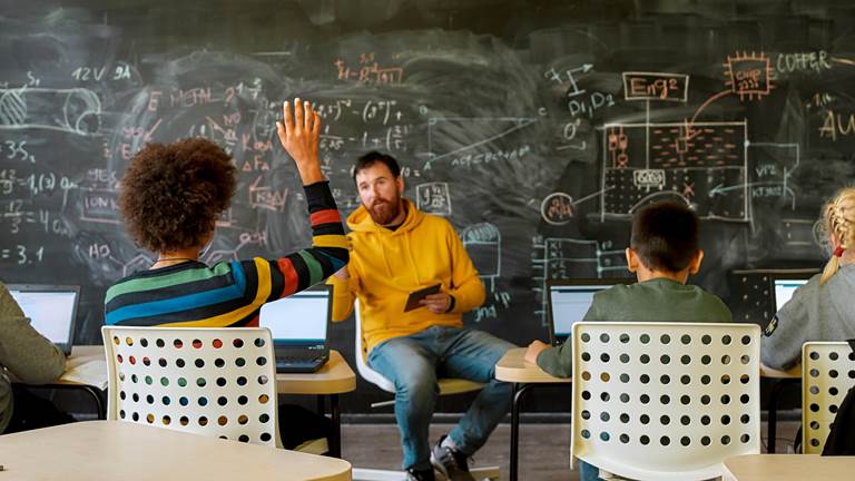 A teacher sitting on a chair holding a tablet in the center of a classroom. A row of students are facing the teacher and are working computer devices. A student with a striped sweater has their hand raised. 