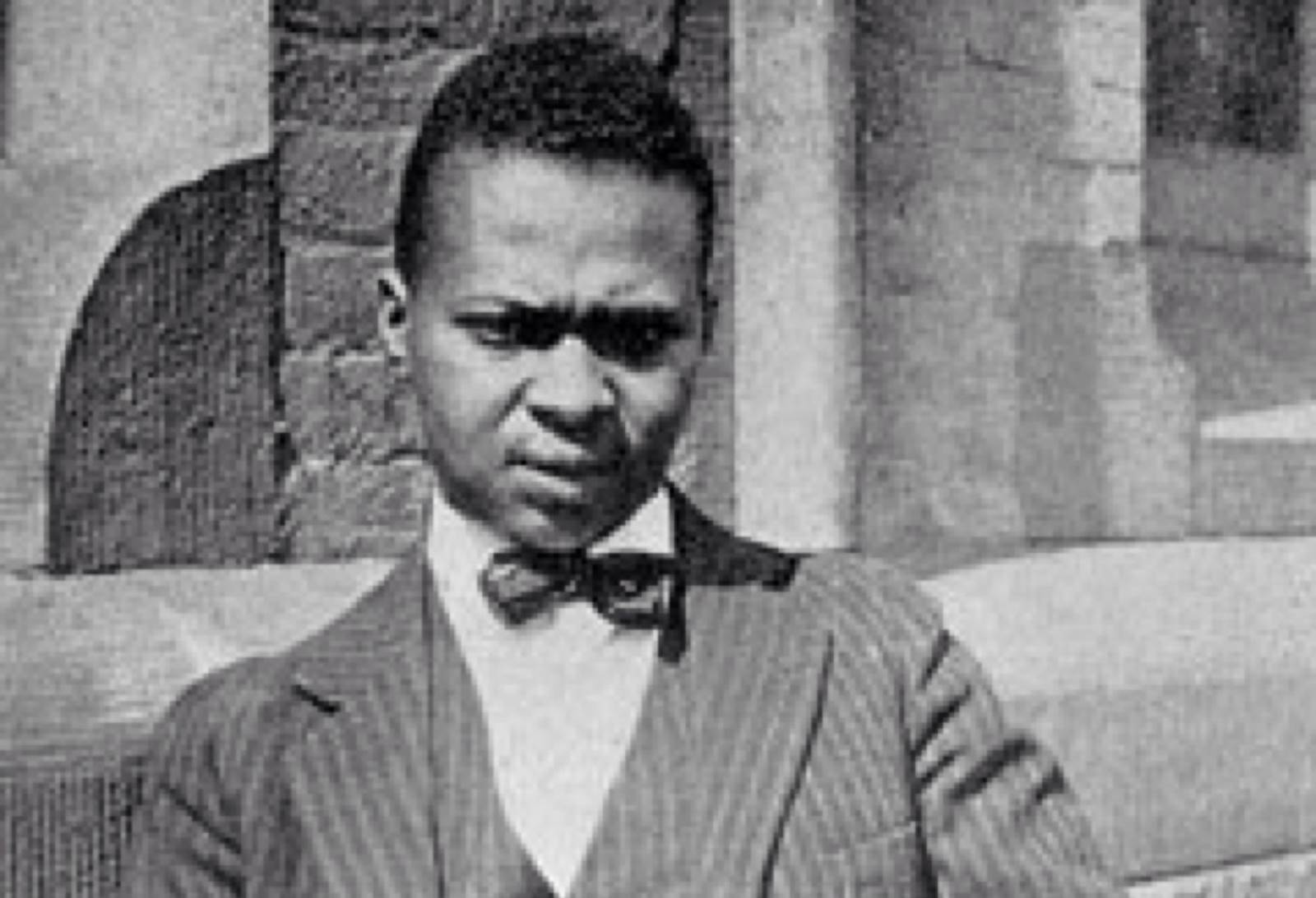 A black-and-white photo of poet and editor Countee Cullen standing next to a brick building. He wears a striped business jacket with a matching vest, a light-colored dress shirt, and dark bowtie.  