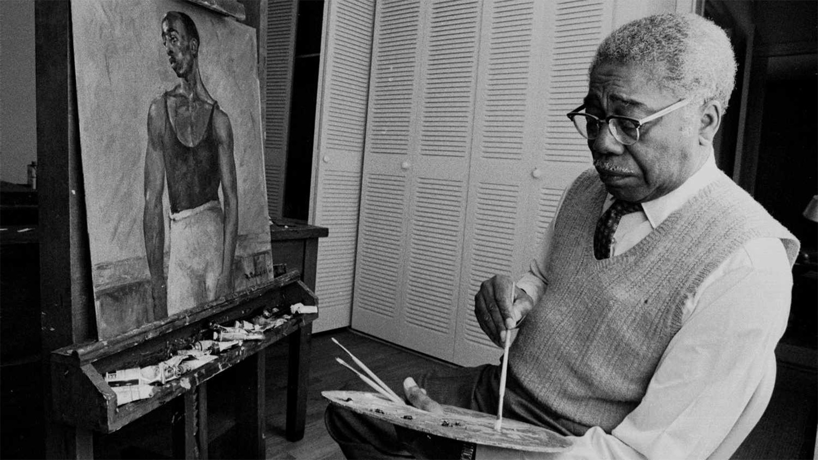 A black-and-white photo of an older Black American man sitting in front of an easel on which there is a painting of a younger Black man in a dark tank top and dancer tights. The artist wears reading glasses, a light-colored sweater vest over a white dress shirt and dark tie, and dark dress pants. He is dipping a paint brush into paint on a palette he holds in his left hand.