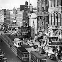 A black-and-white photo of a busy street in Harlem during the Harlem Renaissance period. The photo&rsquo;s perspective is from a high point directed across the street to capture trolleys and pedestrians.