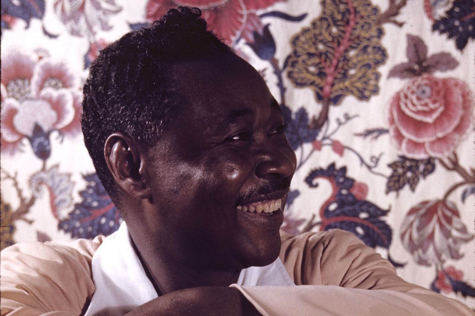 A photo of poet and novelist Claude McKay as he smiles and looks off to the right. He wears a tan shirt with a white collar and is in front of a cream wallpaper with green and pink flowers and leaves.