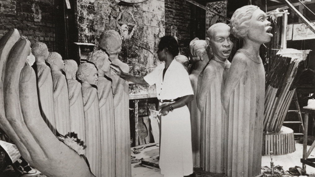 A black-and-white image of Augusta Savage working on a sculpture in her studio. Several human figures in uniform robes pose with their arms behind their backs in a line, mouths open in a singing gesture. A large, curved hand sculpture is on the left side of the photo.