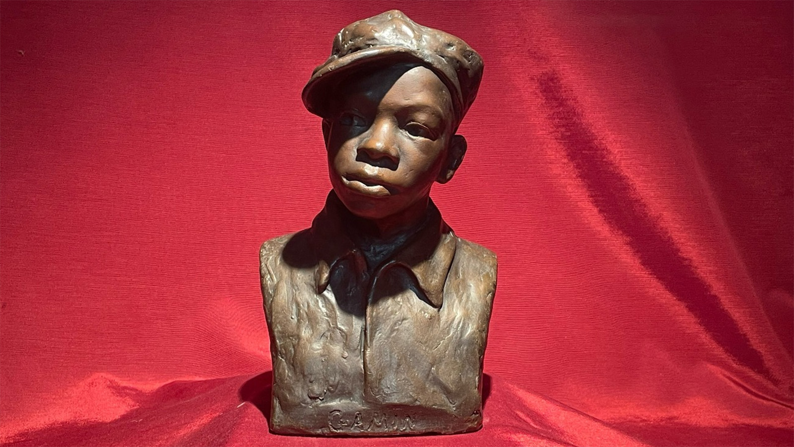 An image of a brown plaster bust of sculptor Augusta Savage&rsquo;s nephew, Ellis Ford. The bust portrays a boy wearing a collared top and a newsboy cap. The bust rests against a red satin backdrop.