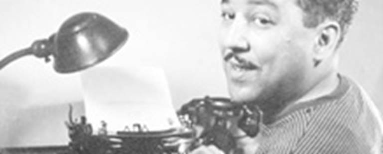 A photo of a Langston Hughes sitting at a desk with his hands posed on a typewriter. 
