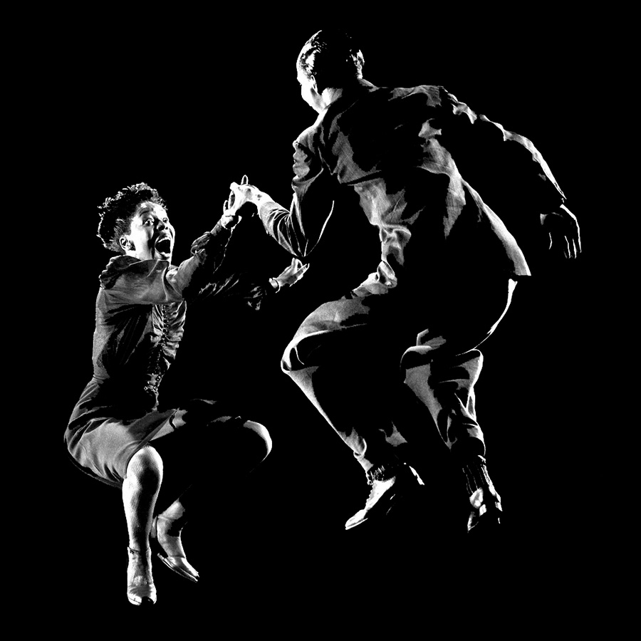 A black-and-white image of two dancers in the air with their knees lifted up to their hips. Both dancers wear dark outfits and move against a black background, illuminated by lighting. The dancer on the left&rsquo;s right hand is in the dancer on the right&rsquo;s left hand.