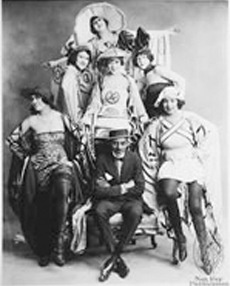 A black-and-white photo of a group of smiling Black performers in 1920s dresses and suits pose for the camera. A performer in a dark suit, dark shoes, white dress shirt, dark bowtie, and hat sits in an armchair with their arms crossed and their legs crossed at the ankles. Two performers perch on the chair&rsquo;s armrests on either side with their hands on their hips. Four performers are posed behind the chair on an elevated platform; one of the four wears a wide-brimmed hat and holds their arms out wide.