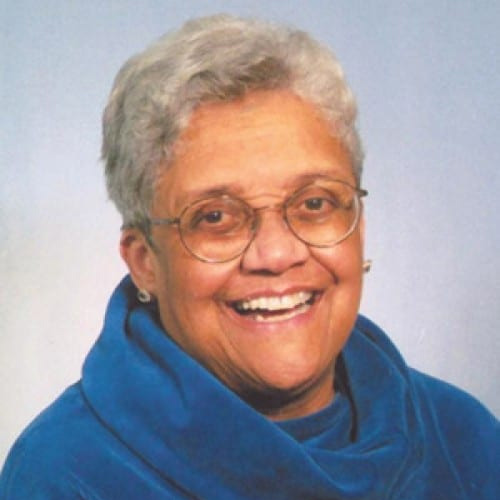 A photo of a smiling Black woman with short silver and white hair smiling. She wears a blue turtleneck top, circular glasses, and earrings.