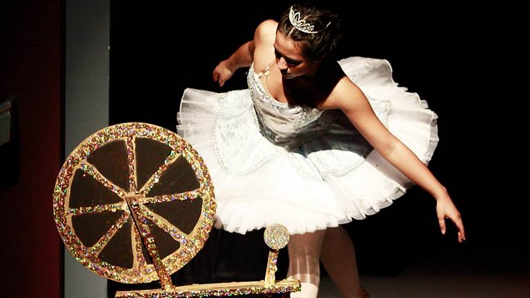 A scene from the play, Sleeping Beauty. Princess Aurora is positioned next to a spinning wheel, looking at it curiously. The wooden spinning wheel is covered in jewels and the ballerina is wearing a white tutu and crown. 