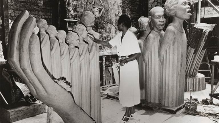 A black and white photograph of Augusta Savage and her statue, The Harp. She is wearing a white dress and is standing in between two parts of the statue that stand taller than her. The Harp is made of 12 black singers standing in graduated heights to symbolize the strings of a harp. The sounding board is the hand and arm of God.