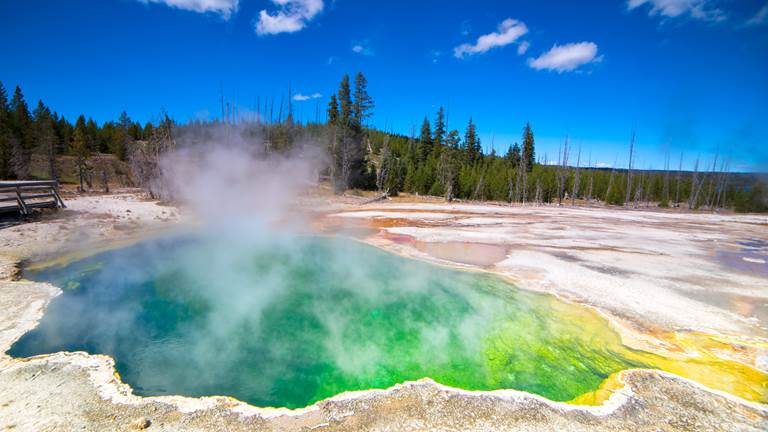 A photograph of the Yellowstone Caldera. The water is blueish-green and there is steam rising up.