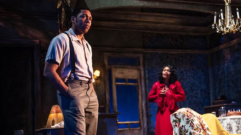 A scene from "A Streetcar Named Desire." A man is in the foreground with his hands in his pant pockets. A woman in a red dress standing in the background with her hands clasped against her chest.