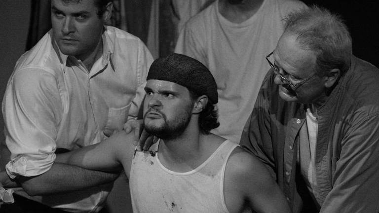 A scene from the play The Hairy Ape. A man is in the center of the photo. He is sweating with an angry look on his face while two men hold him down by his arms.