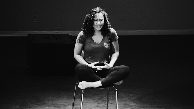An actor on stage sitting in a chair with their legs criss crossed.  