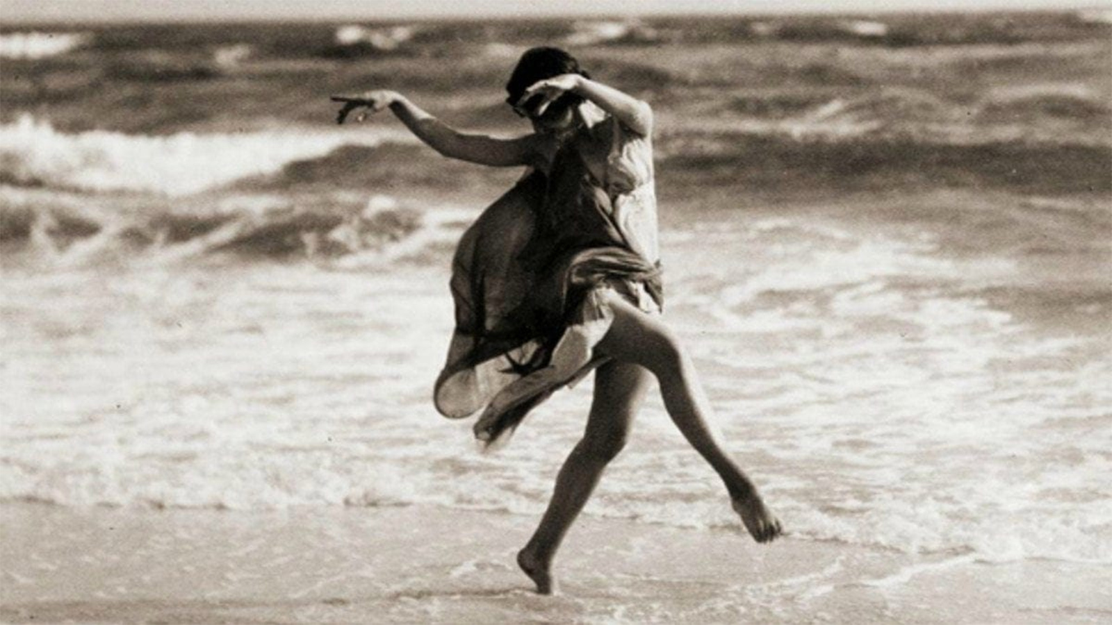 A vintage photo of a woman dancing along the edge of a beach. She dances barefoot in the sand and wears a flowy dress.