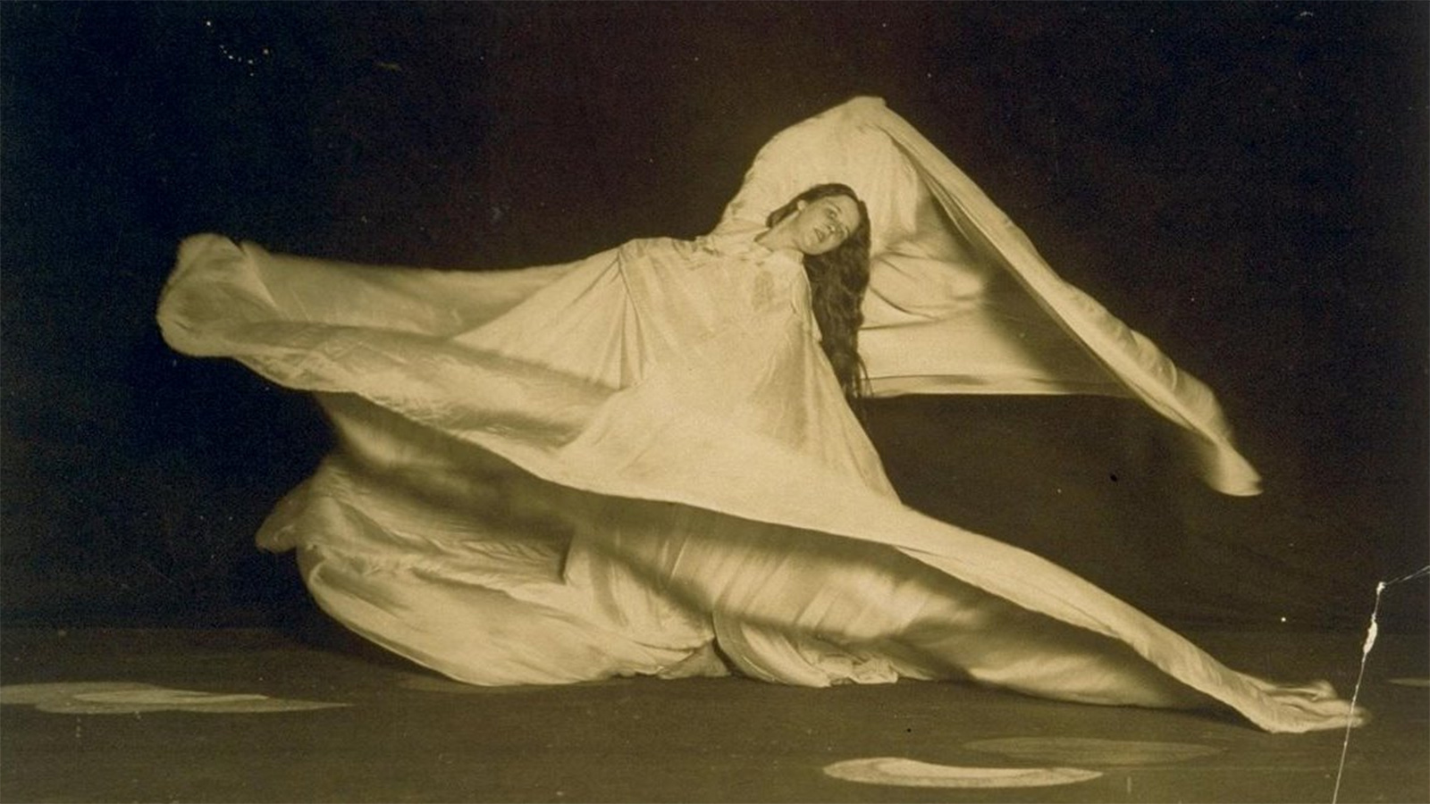 A vintage photo of a woman with long dark hair dancing while wearing a light-colored costume with dramatically long sleeves that swirl in motion.