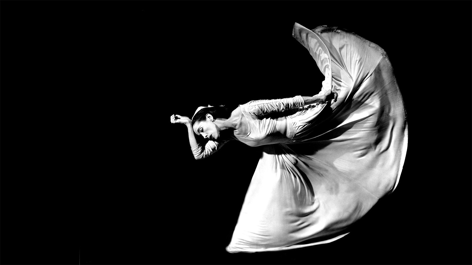 A black and white photo of a female dancer in a dramatically lit pose. Her torso is parallel to the ground with her right wrist placed against her head. Her dress&rsquo;s long skirt billows in an arch shape.
