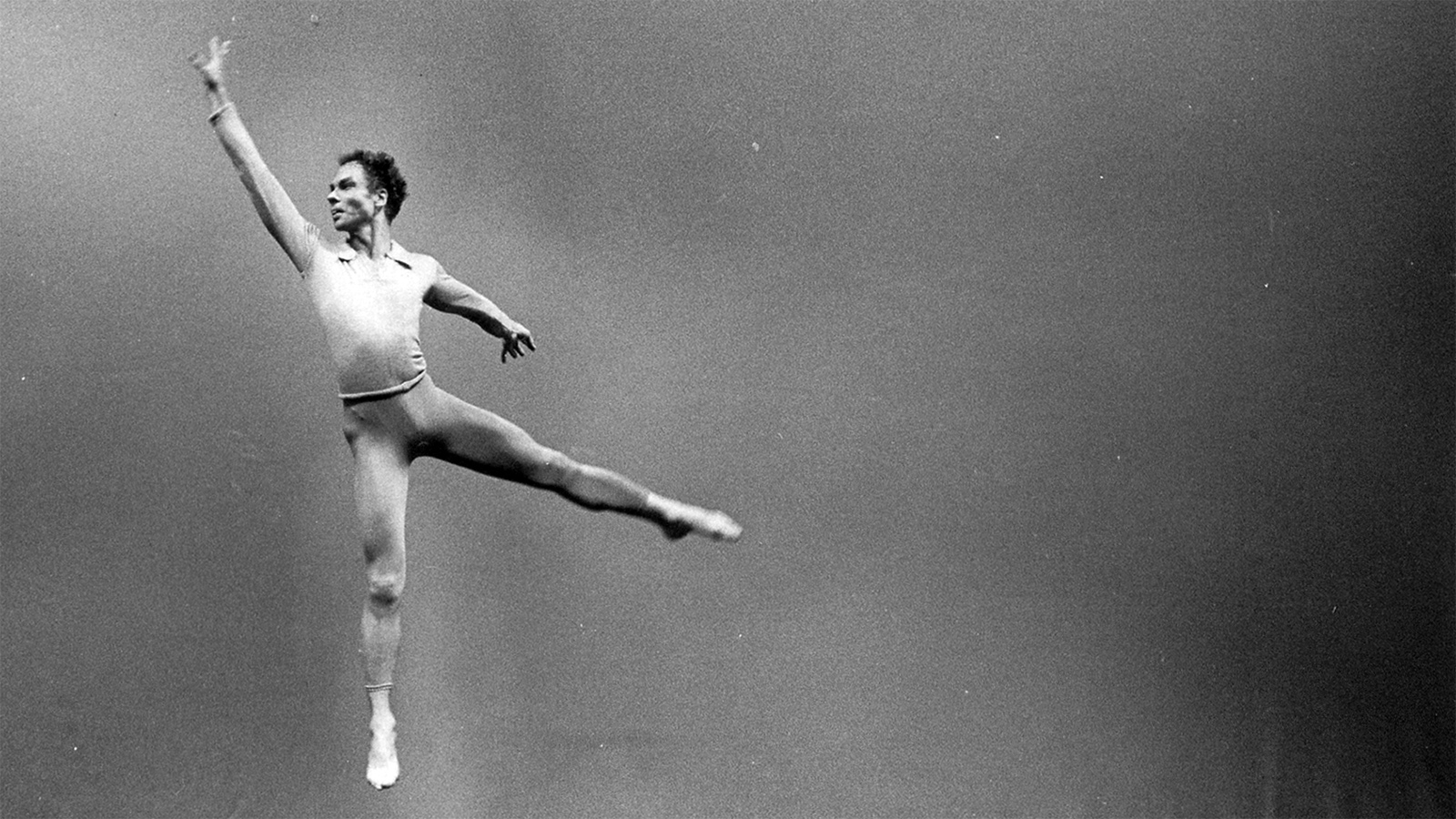 A black and white photo of a male dancer leaping into the air. His right arm is raised into the air while he extends his left arm and left leg away from his body.